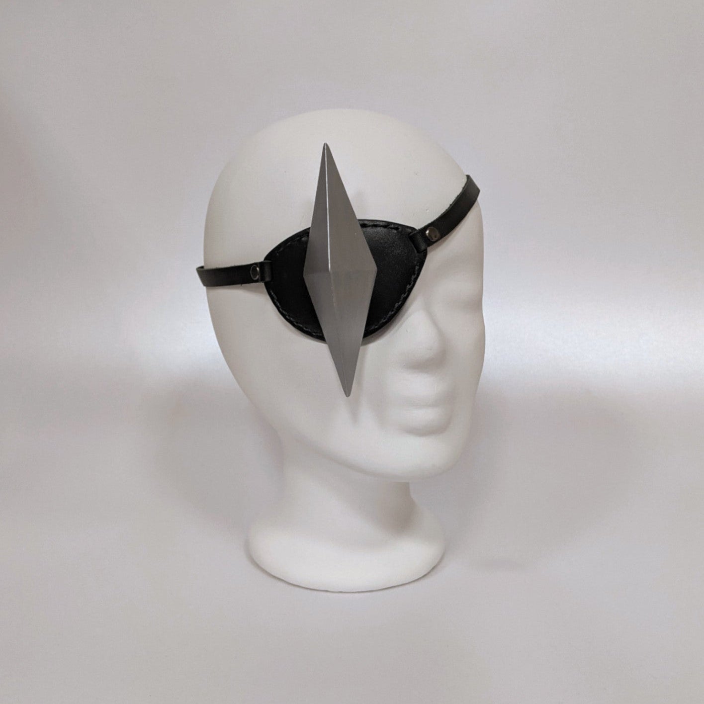 A white mannequin head adorned with an eyepatch with a geometric shape on top, set against a soft light background.