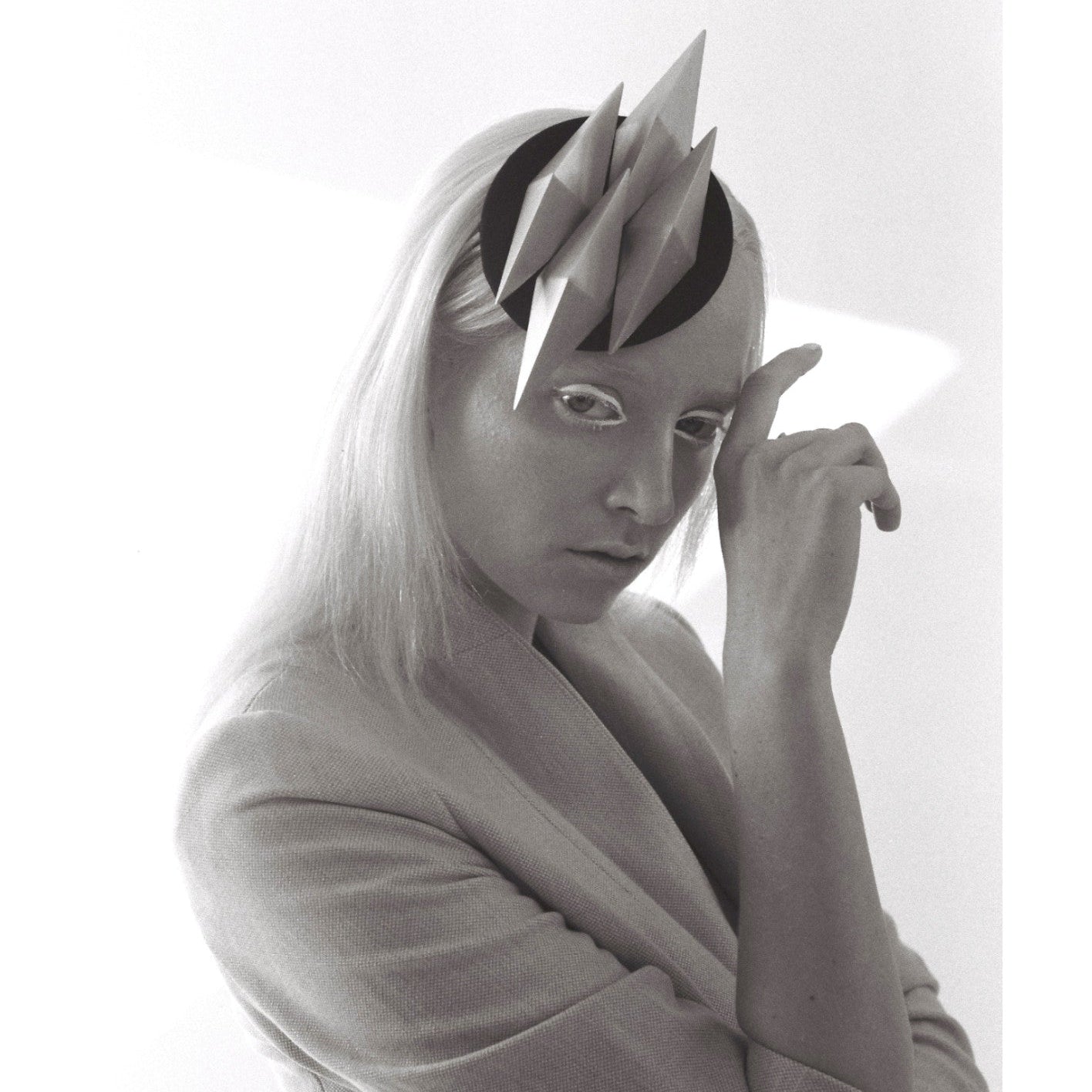 A figure in a grey blazer poses with one hand raised, showcasing an artistic headpiece of angular shapes against a minimalist backdrop.