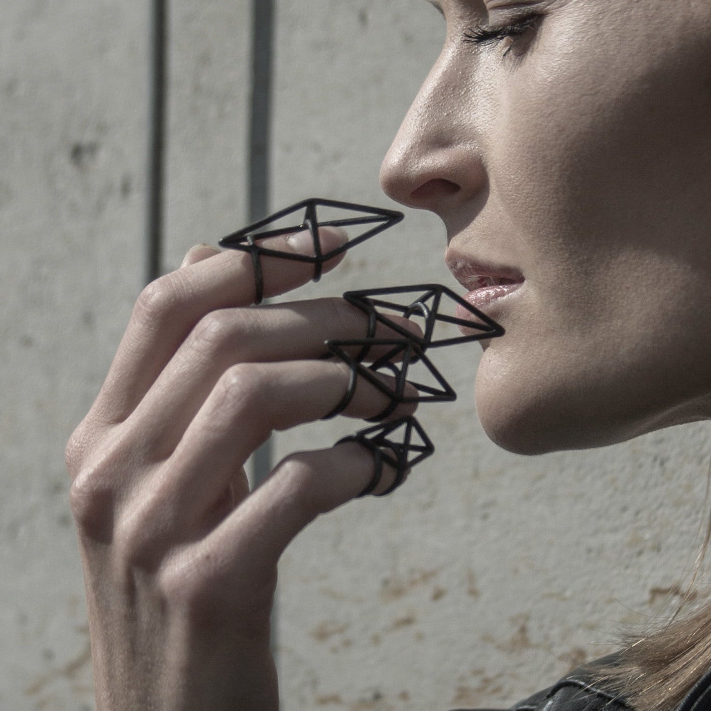 A delicate hand adorned with geometric black wire rings, set against a textured concrete wall, showcasing a trend-forward accessory style.