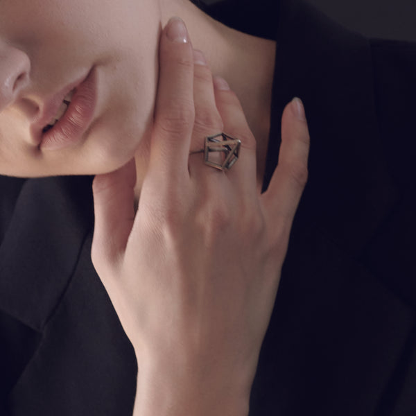 Close-up of a woman's hand touching her chin, wearing a large, geometric ring, with a blurred black jacket background.