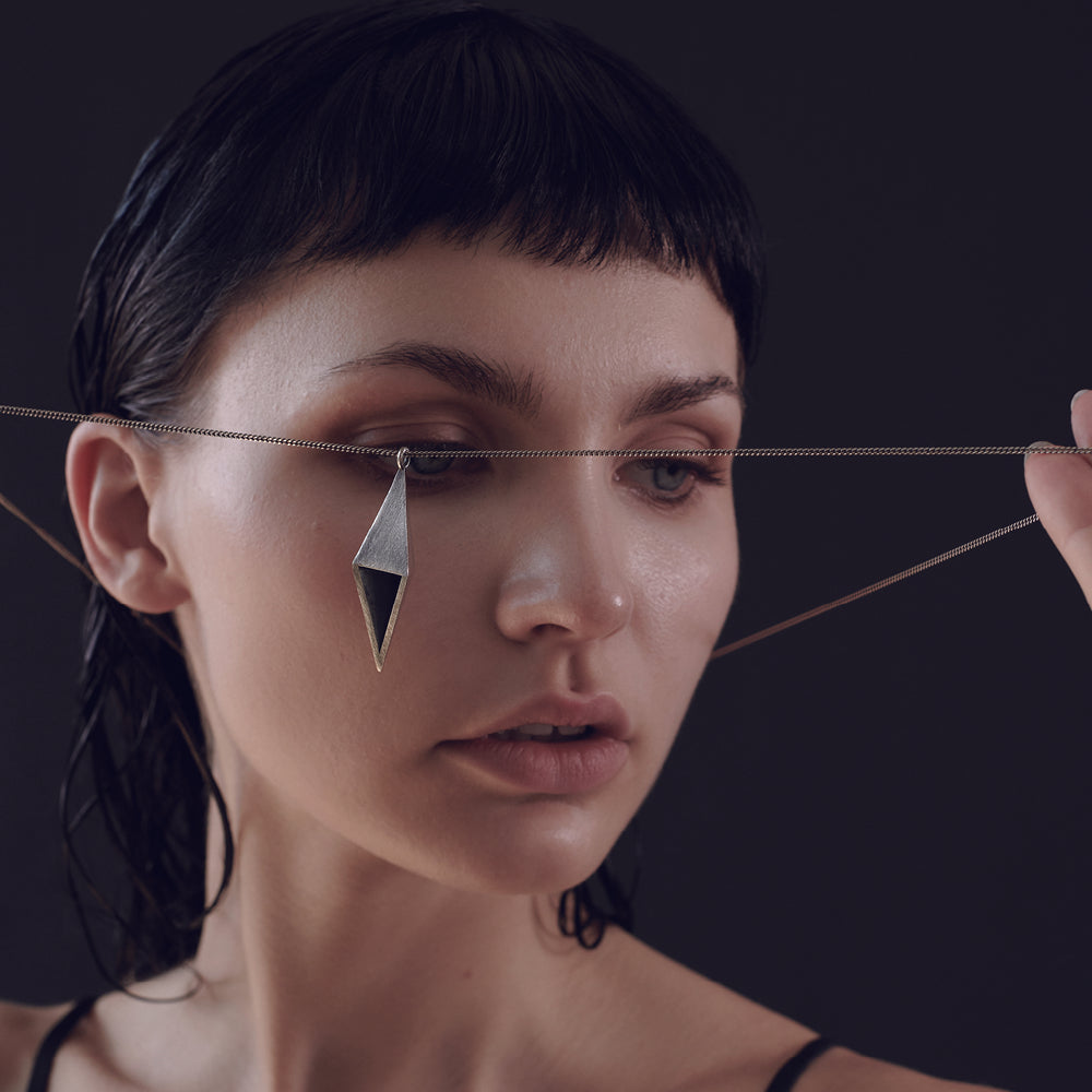 A person with wet, dark hair holds a silver chain with a geometric pendant in front of her face.