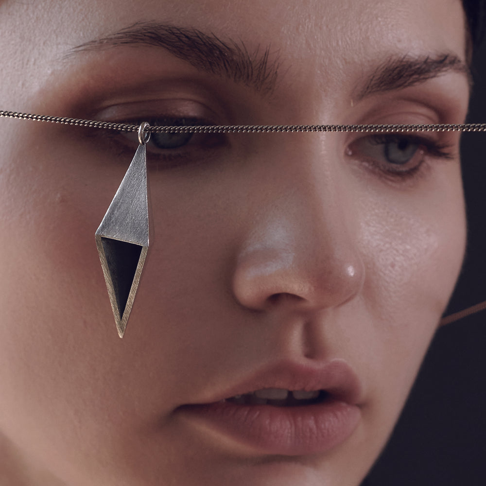 A close-up of a person with wet, dark hair holding a silver chain with a geometric pendant in front of her face.