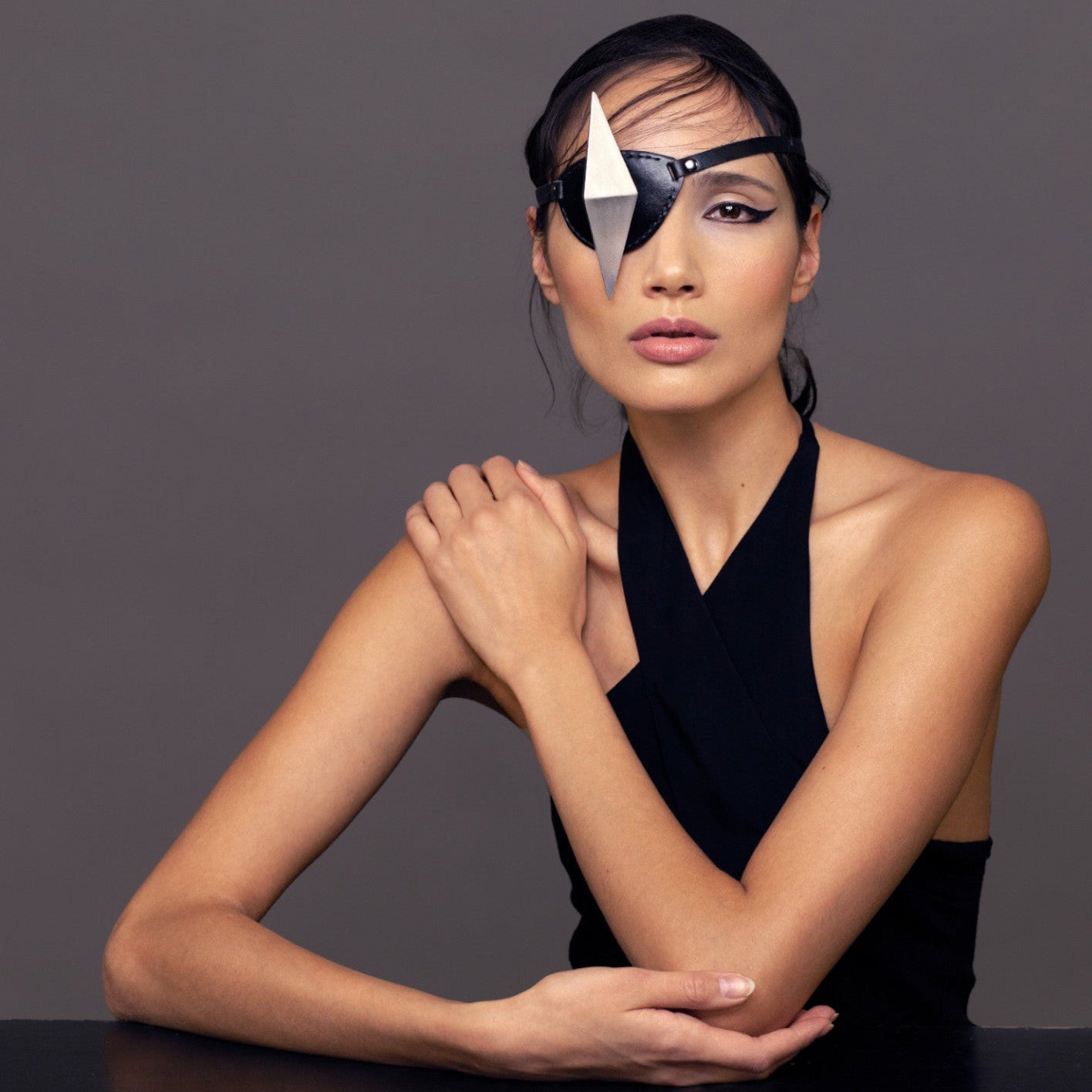A close-up portrait of a woman with dark hair, wearing a black halter top and eyepatch with a silver geometric shape, gently resting her hand on her shoulder against a gray background.