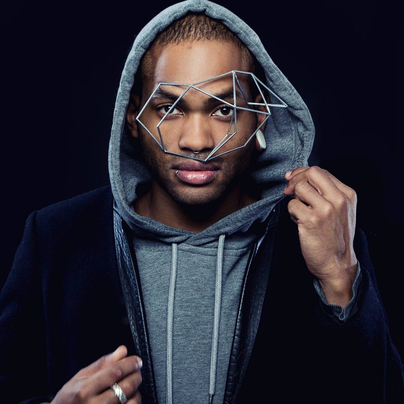 A person wearing a grey hoodie under a black jacket, standing against a dark background, showcasing casual urban style.