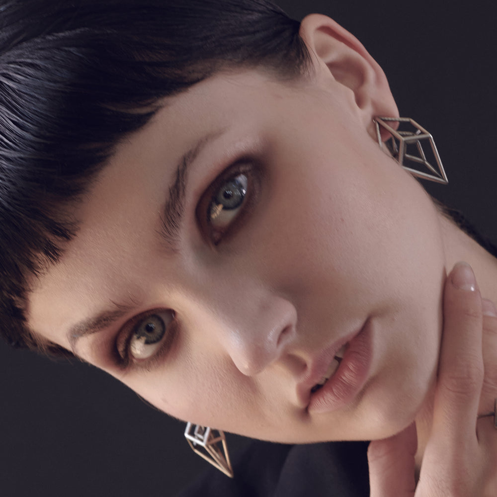 A person with sleek dark hair and wearing a stylish outfit and silver geometric earrings, holding their hand near their face against a dark background.