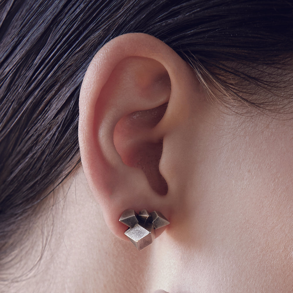 Close-up of a human ear with smooth skin and dark, straight hair in the background. The ear showcases its natural curves and contours and is wearing a silver geometric earring.