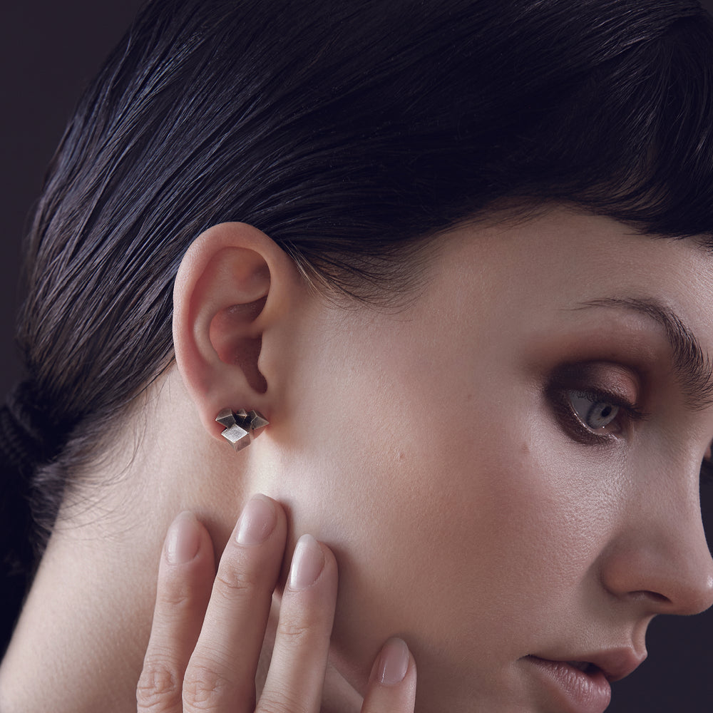 Close-up of a hand gently touching the earlobe, showcasing a geometric silver earring against a dark background.