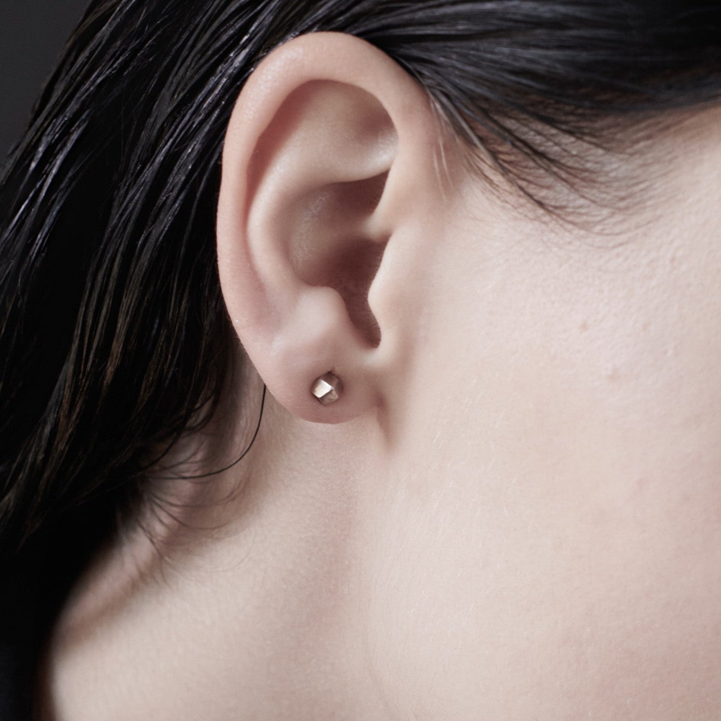Close-up of a woman's ear showcasing a small, minimalist geometric stud earring against sleek, dark hair.