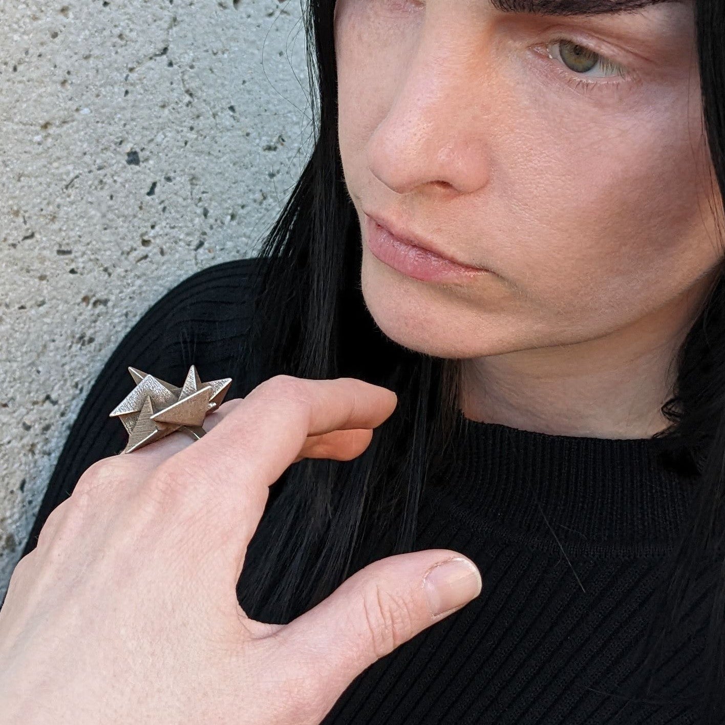 A person wearing a black shirt, showcasing a stylish, edgy ring on their hand against a cement background.