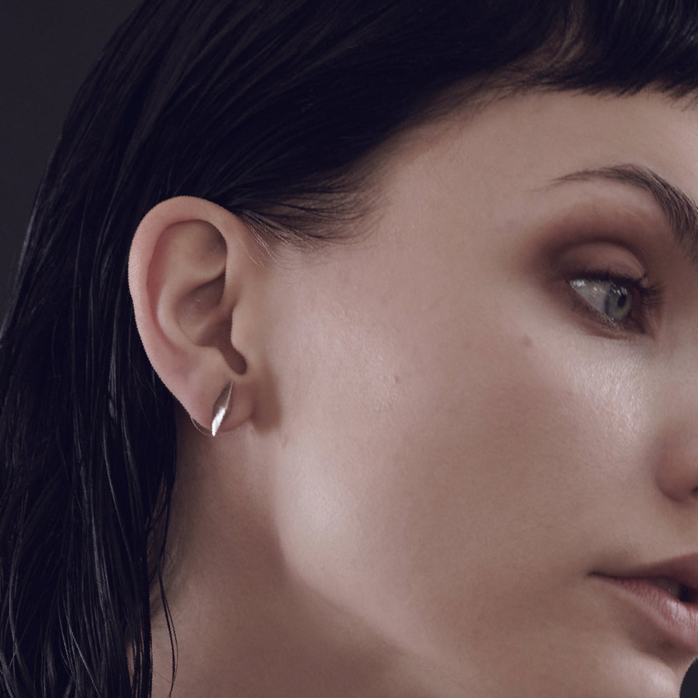 A close-up of a woman's ear adorned with a sleek, silver earring, showcasing dark, wet hair and a minimalistic style against a dark background.