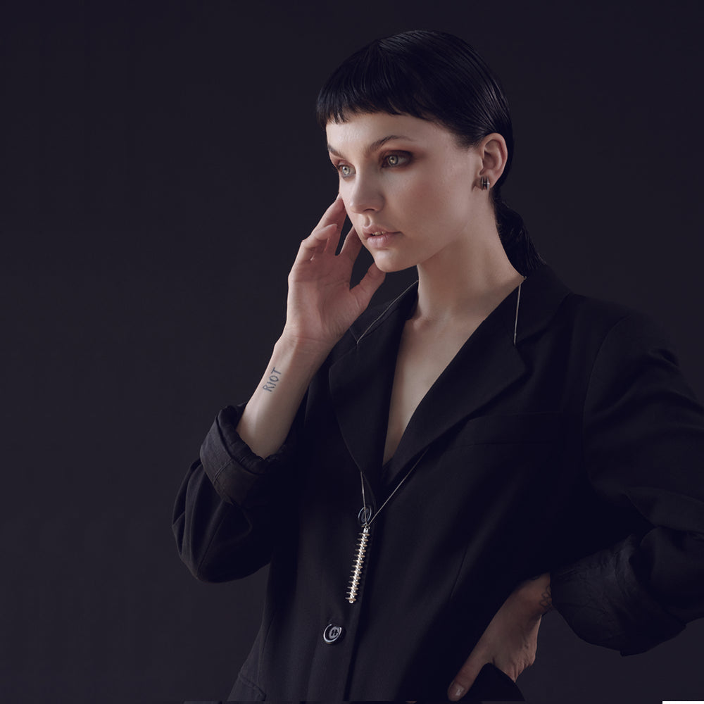 A person in a stylish black blazer with a long silver necklace poses against a dark background, showcasing minimalist fashion.