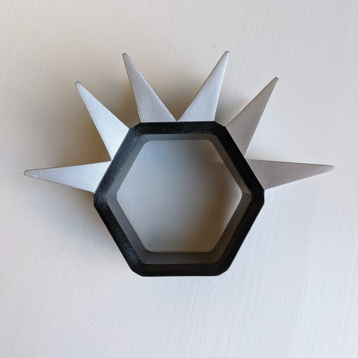 A hexagonal object with a black center and six metallic silver spikes radiating outward, set against a light background.