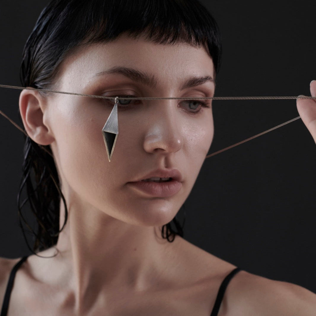 A person with wet hair holds a thin chain with a geometric pendant close to their ear, set against a dark background.