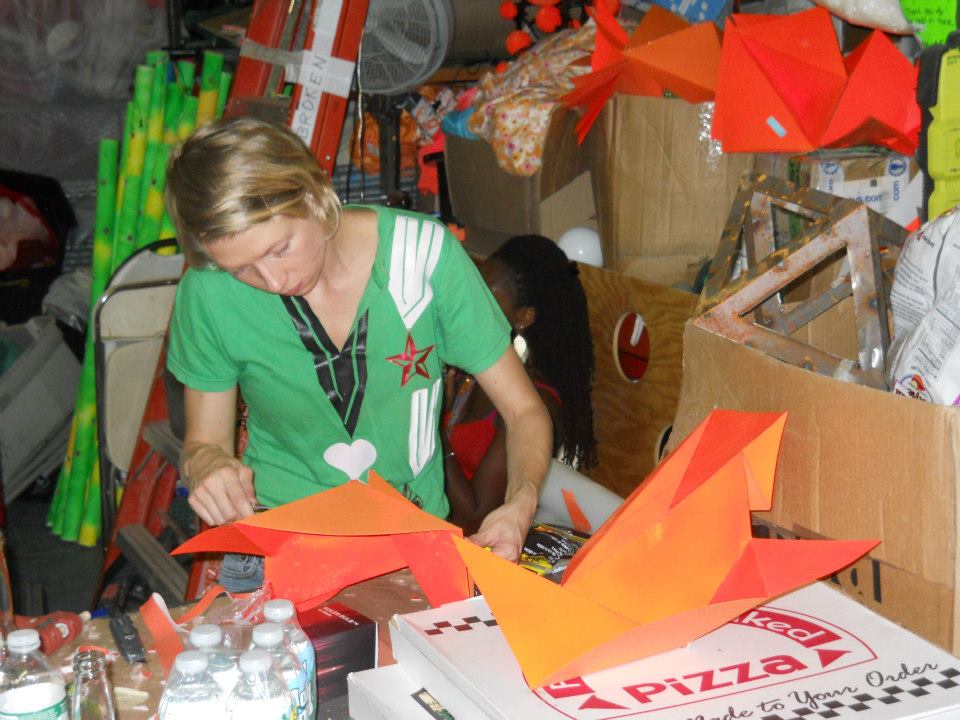 A person assembles brightly colored orange paper shapes in a cluttered workshop filled with craft supplies and tools.