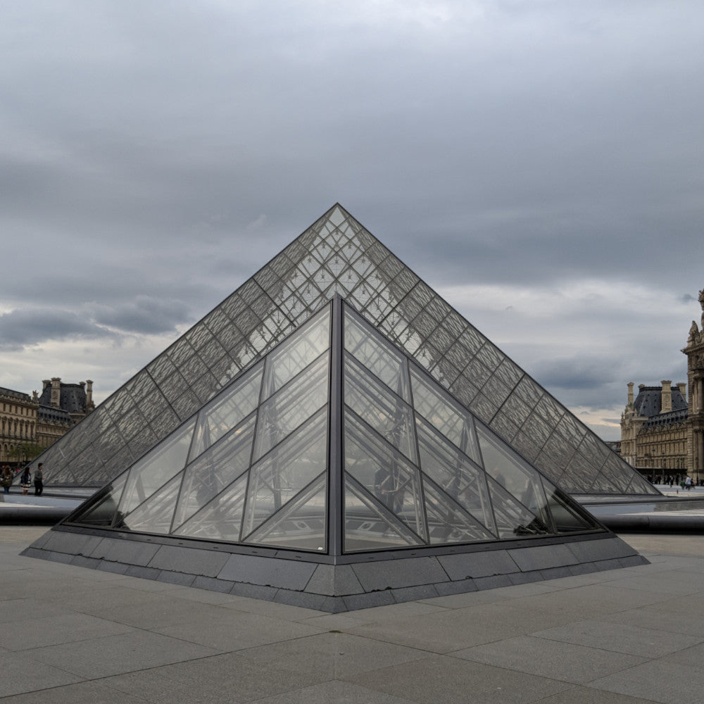 The glass pyramid of the Louvre Museum rises against a cloudy sky, surrounded by visitors admiring its modern architecture.