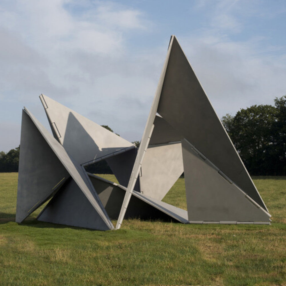An abstract concrete sculpture with angular, overlapping triangular shapes, set in a grassy field under a cloudy sky.