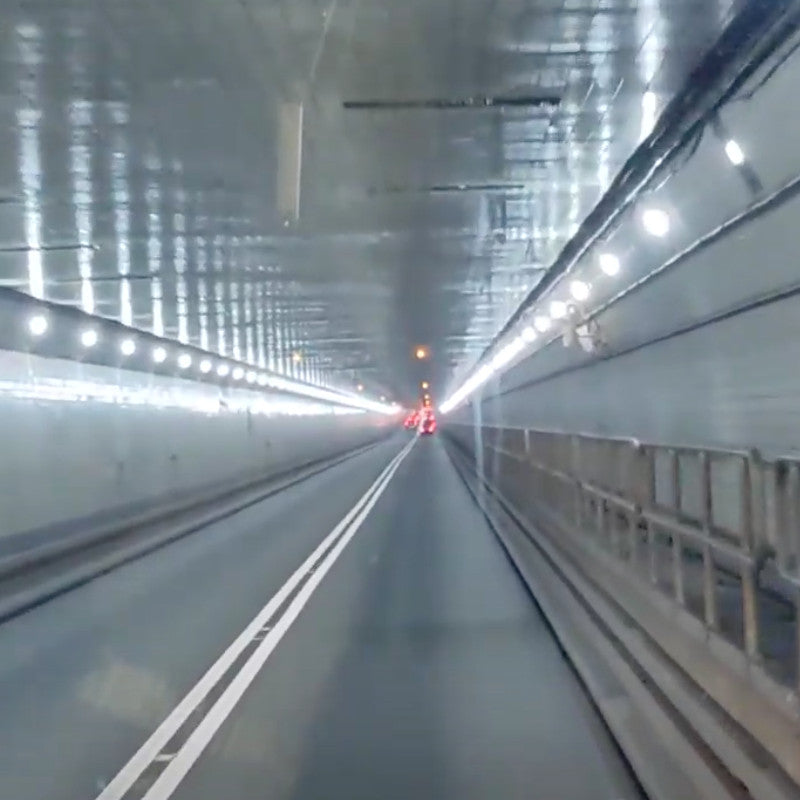 A view inside a well-lit tunnel with a smooth asphalt road, featuring lane markings and approaching vehicle headlights in the distance.