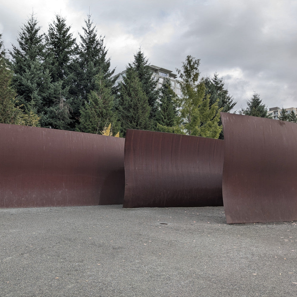 Three large pieces of wavy steel sitting on a grey ground in front of trees and a cloudy sky 