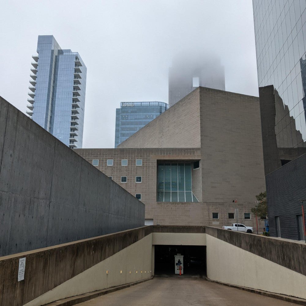 A foggy urban scene features a modern museum entrance, flanked by tall buildings, with mist obscuring the upper parts of the structures.