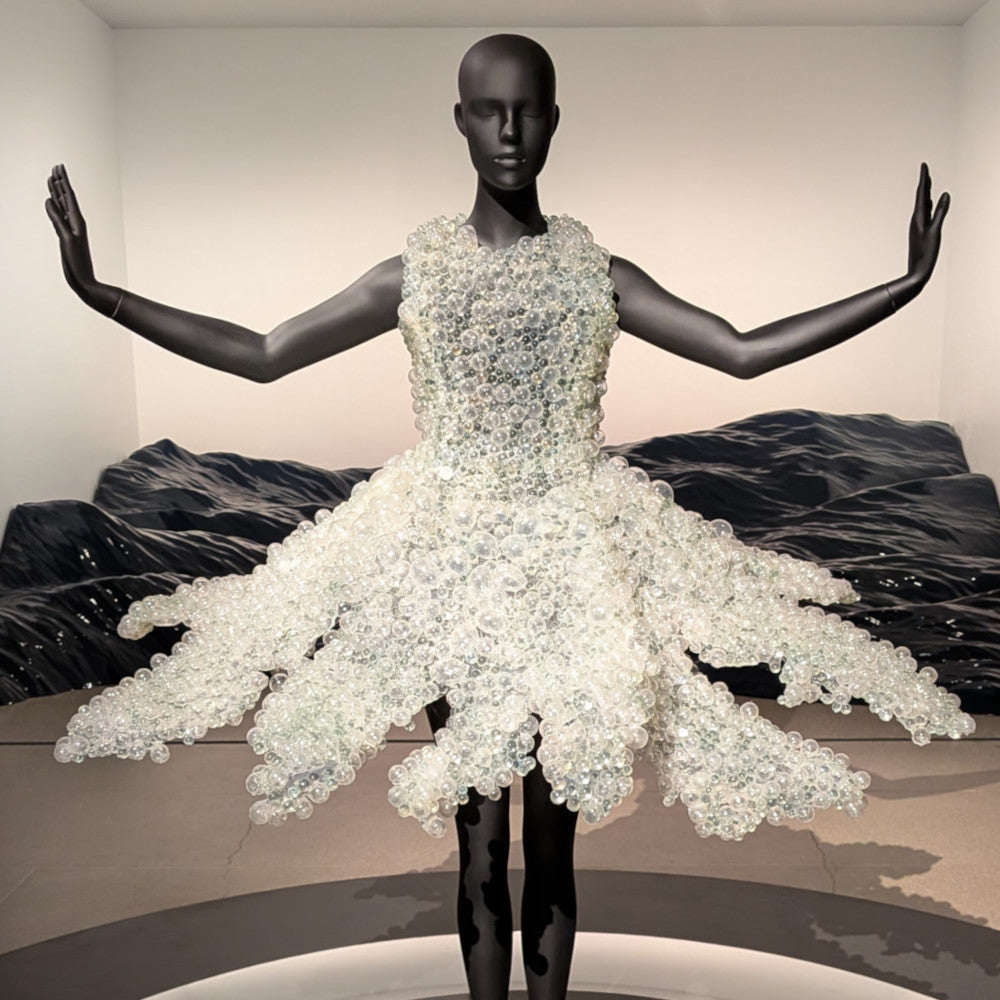 Black mannequin wearing a dress made of numerous clear, translucent beads, with arms extended, displayed against a neutral background.