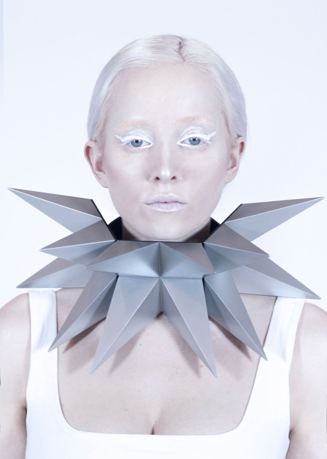 A model wears a striking geometric silver collar made of sharp, star-like folded shapes, against a minimalist white background.