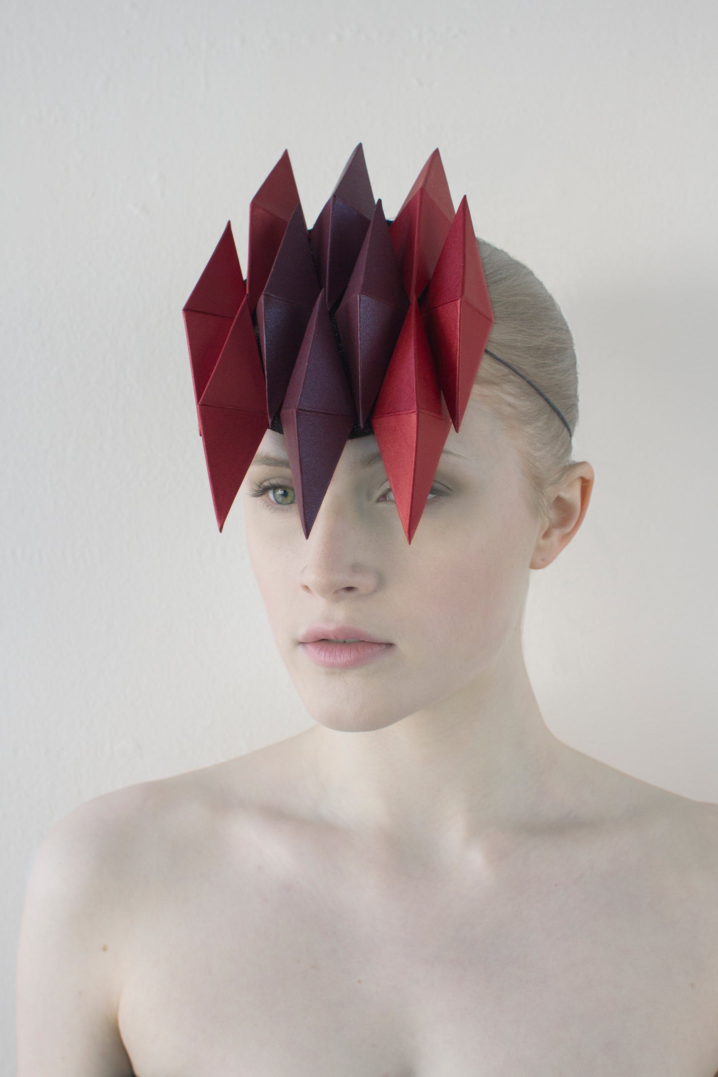 A woman with pale skin in front of a white background wearing a headpiece made of ten purple and red pyramids. 