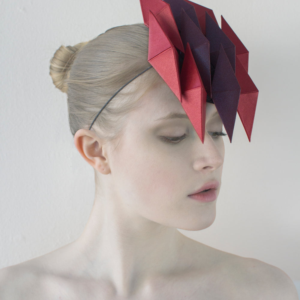 A woman looking to the left with pale skin in front of a white background wearing a headpiece made of ten purple and red pyramids. 