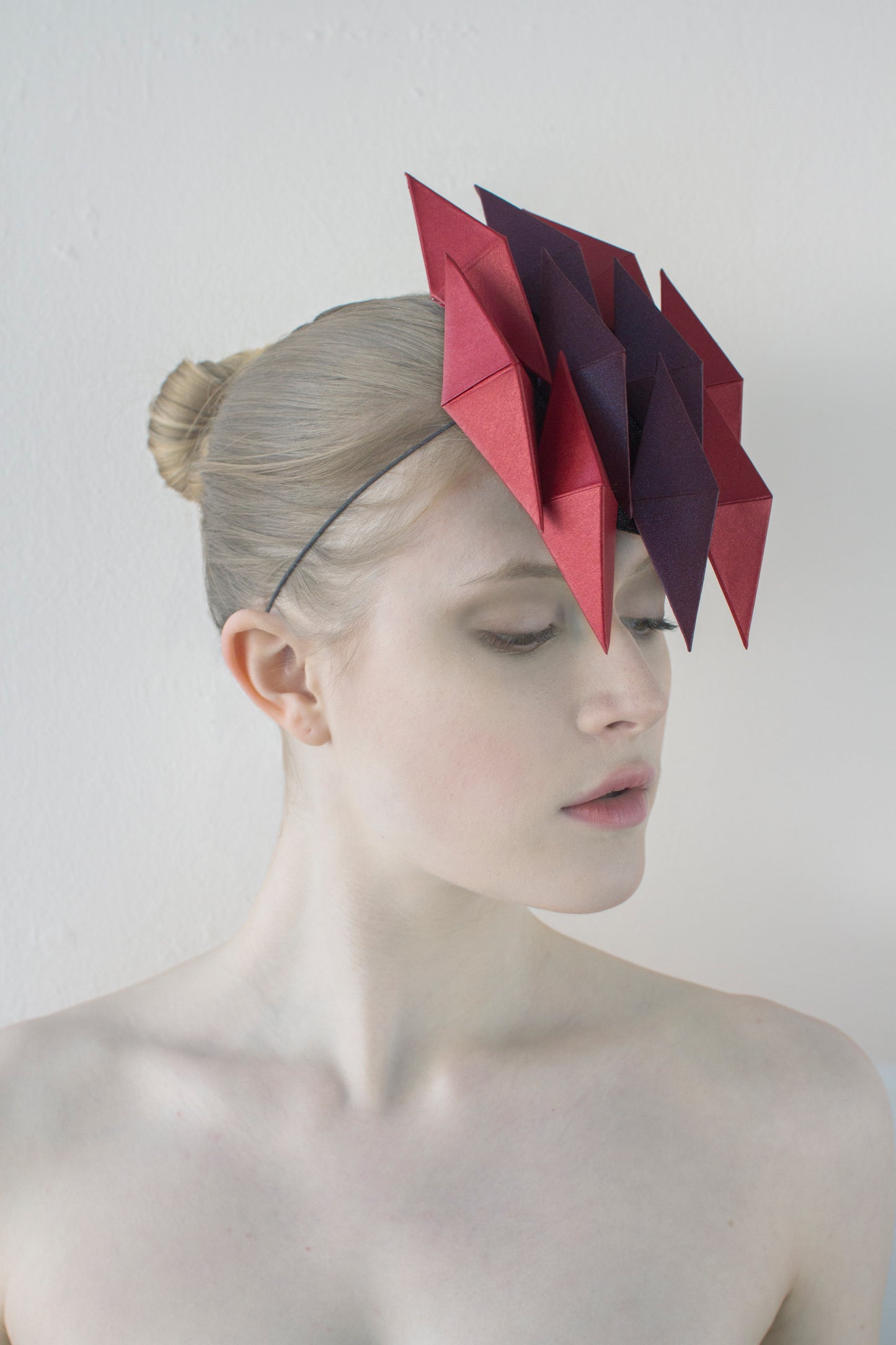 A woman looking to the left with pale skin in front of a white background wearing a headpiece made of ten purple and red pyramids. 