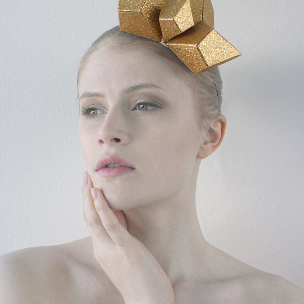 A gold, geometric headpiece made from four pyramids sits on the head of a woman with her right hand touching her chin, sitting in front of a white background