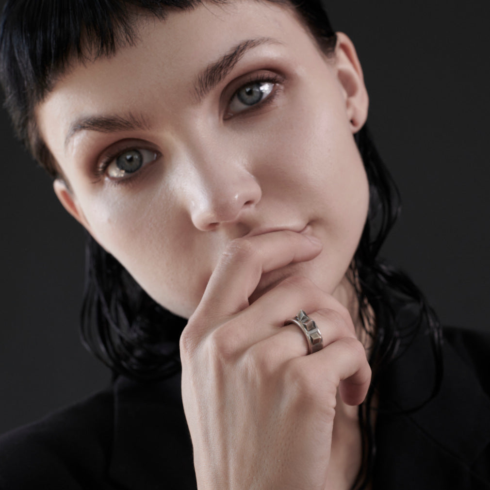 A close-up portrait of a woman with short dark hair wearing a silver ring with dark inlay details on her ring finger, set against a dark gray background.