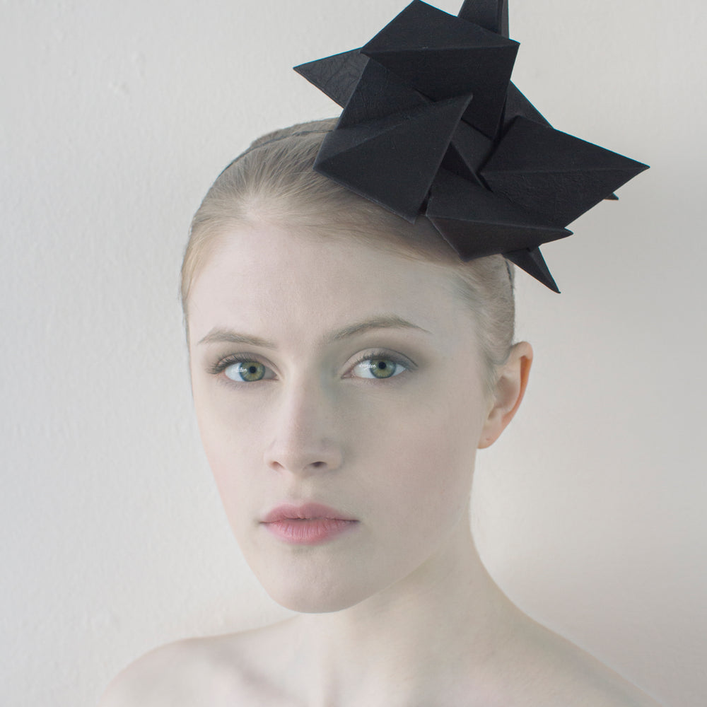 A black geometric headpiece sits on the head of a woman sitting in front of a white background. 