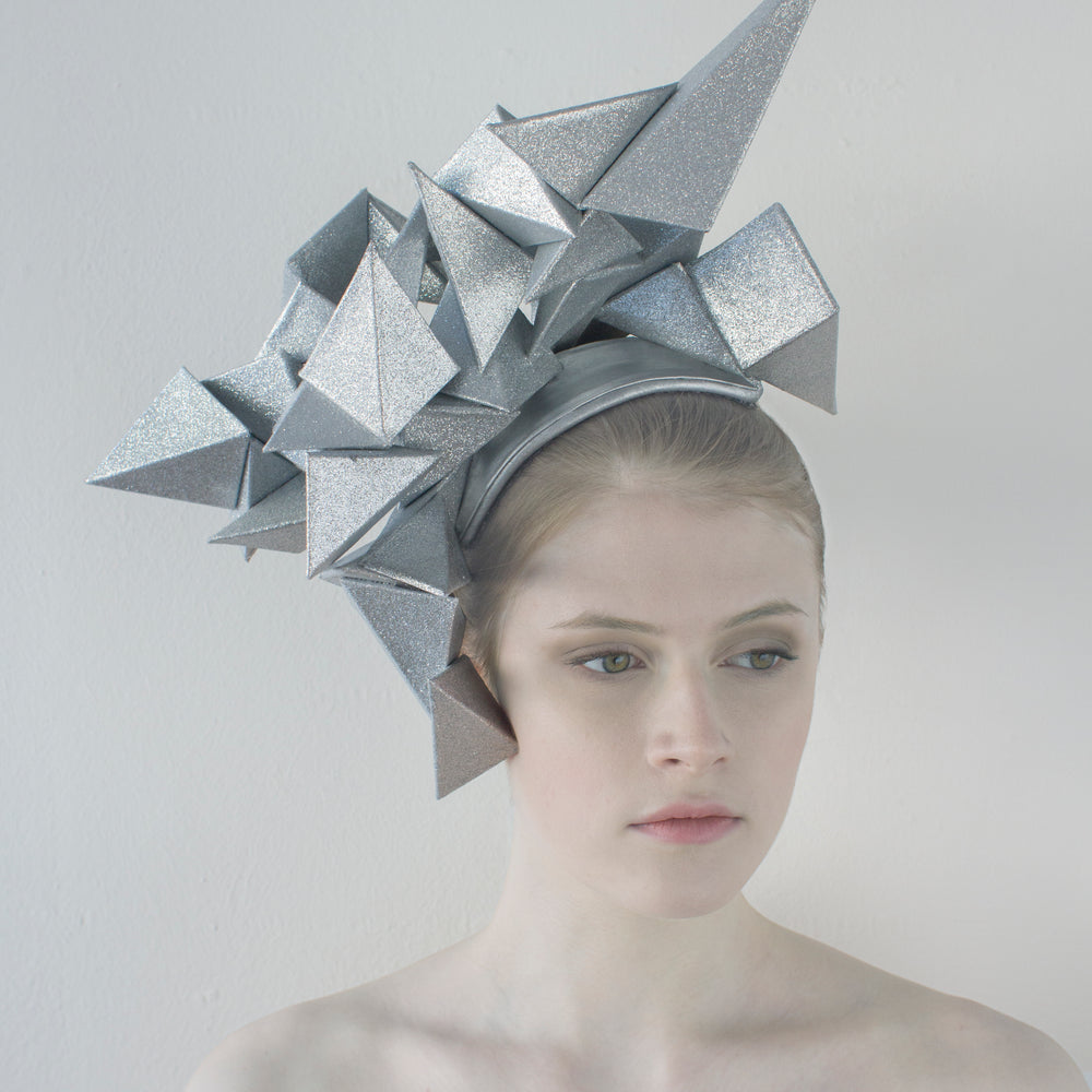 A woman looks slightly to the left and sits in front of a white background wearing a large, silver geometric headpiece that juts out in different directions. 