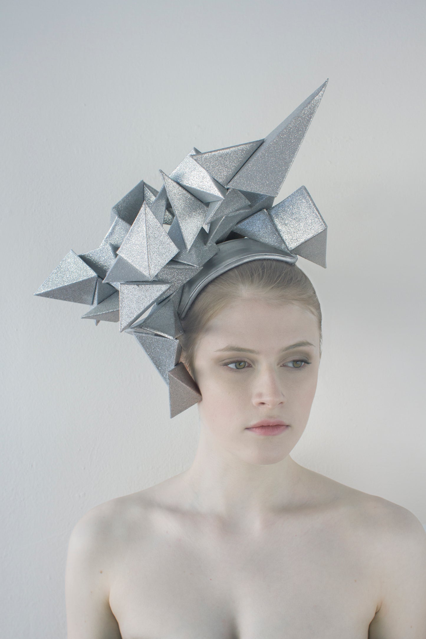 A woman looks slightly to the left and sits in front of a white background wearing a large, silver geometric headpiece that juts out in different directions. 