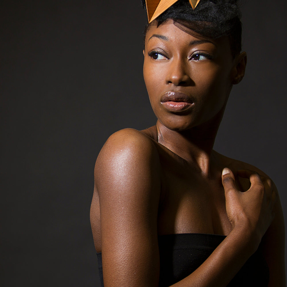 
                  
                    A woman stands in front of a dark background looking over her shoulder, wearing a wood geometric headpiece. 
                  
                