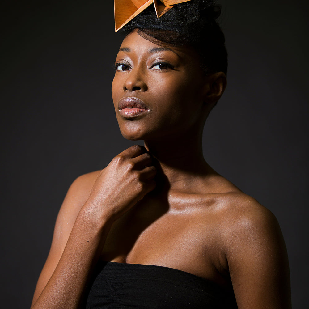 
                  
                    A woman stands in front of a dark background touching her hand to her neck, wearing a wood geometric headpiece. 
                  
                