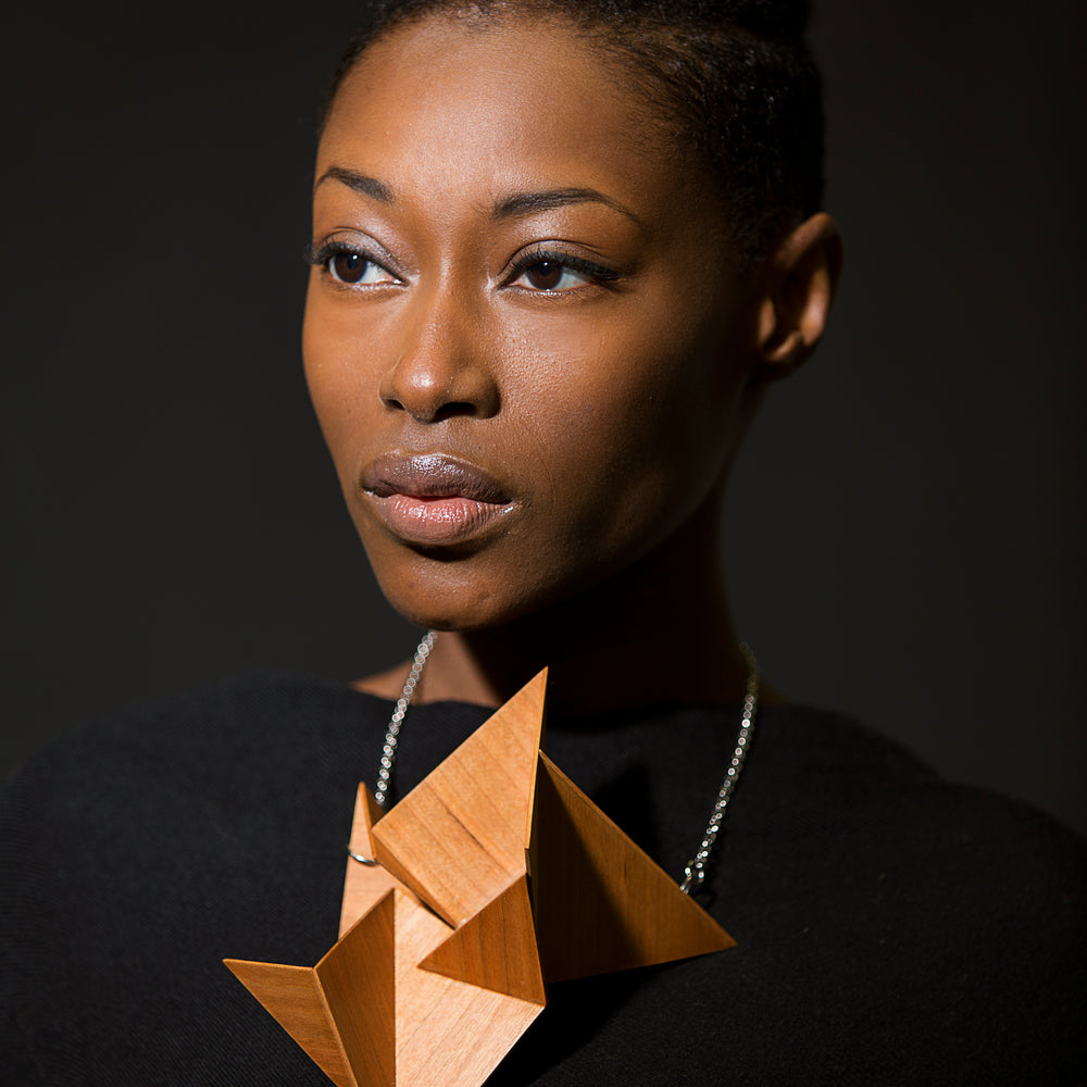 
                  
                    A person wears a large, geometric origami-style wooden necklace against a dark background, showcasing modern artistic jewelry.
                  
                