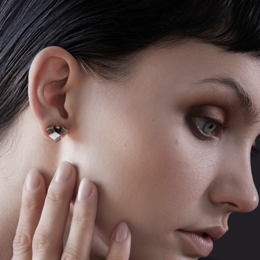 A close-up of a model's ear wearing a small geometric silver stud earring, with her hand resting near her cheek against a dark background.
