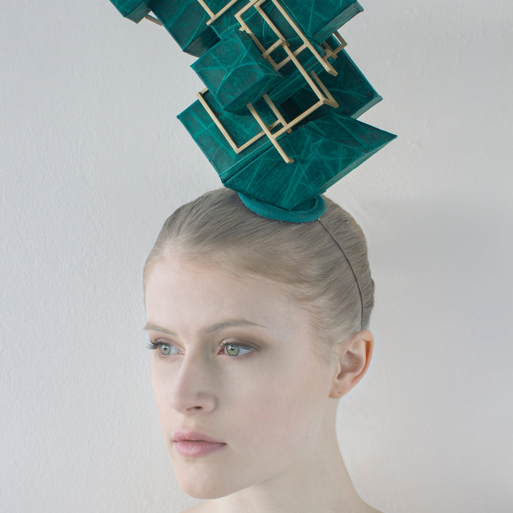 A woman sitting in front of a white background wears a green, sculptural headpiece that has a gold lattice attached to it. 