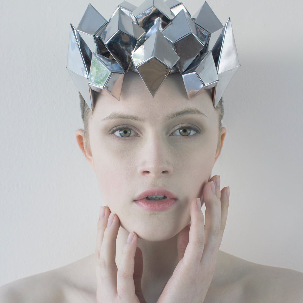 A woman looks straight ahead with both hands touching her chin, and is wearing a silver shiny, geometric crown. 