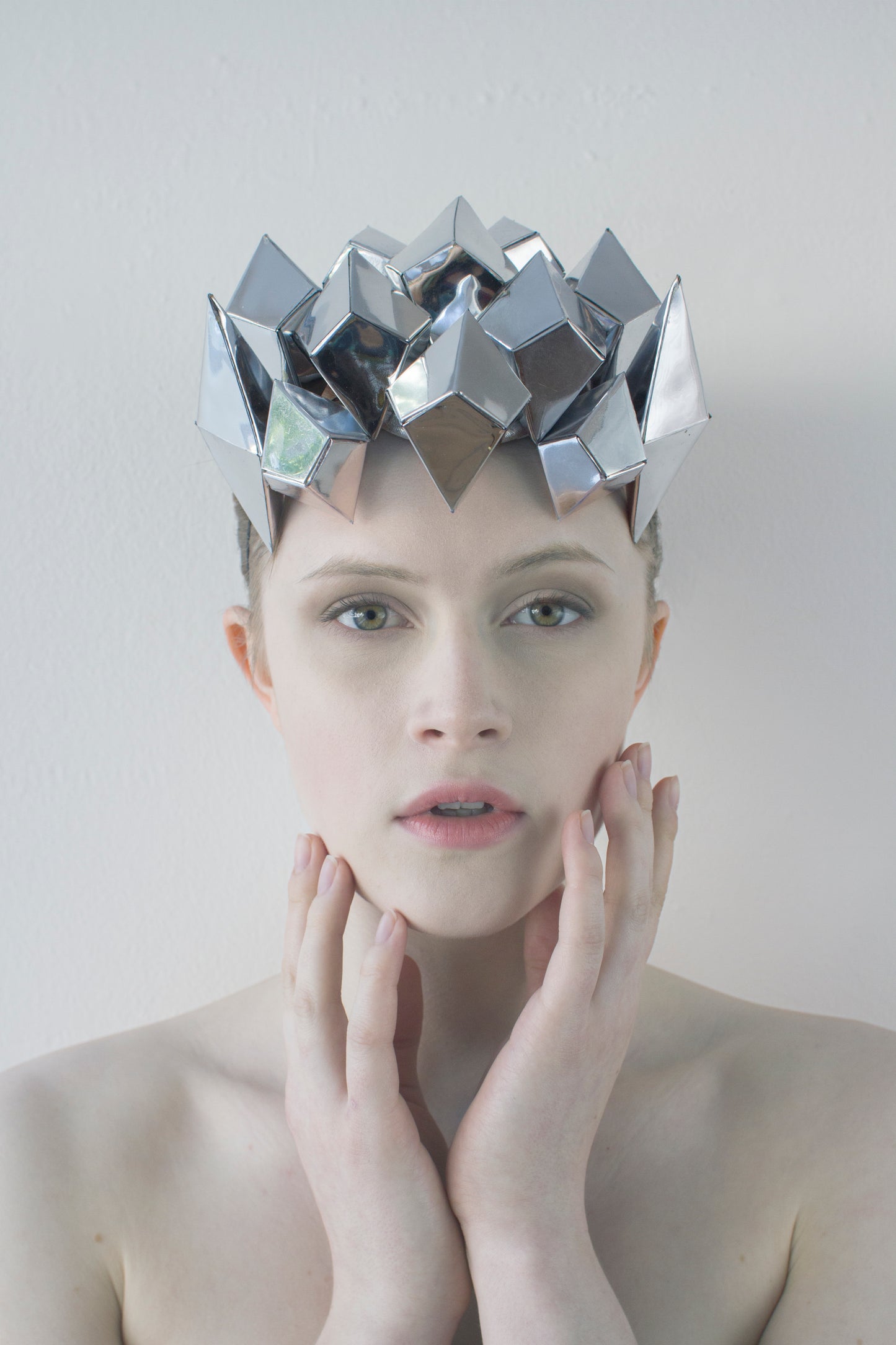 A woman looks straight ahead with both hands touching her chin, and is wearing a silver shiny, geometric crown. 