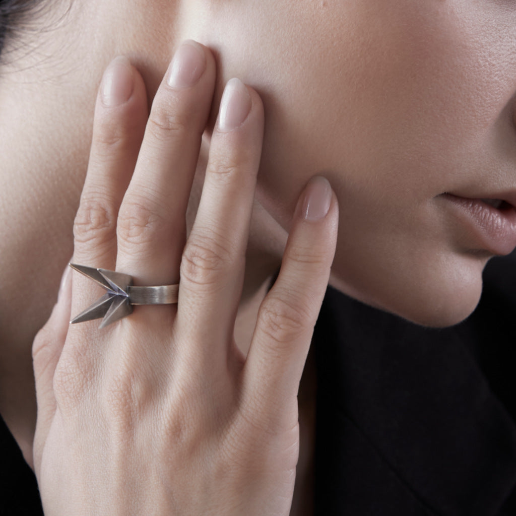 A close-up of a person's hand wearing a silver geometric ring with sharp angular points, resting against their cheek.