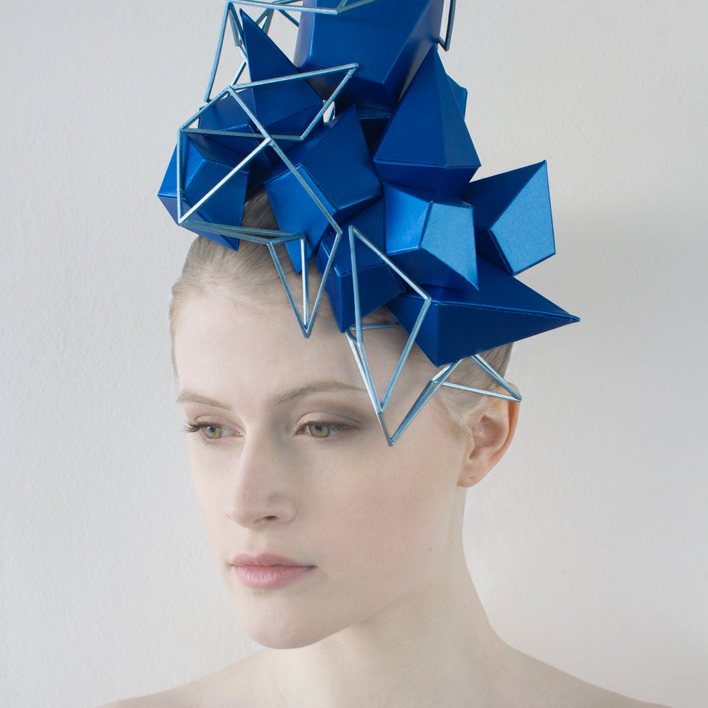 A woman sitting in front of a white background wears a shiny blue, geometric headpiece that is enhanced with a light-blue lattice. 