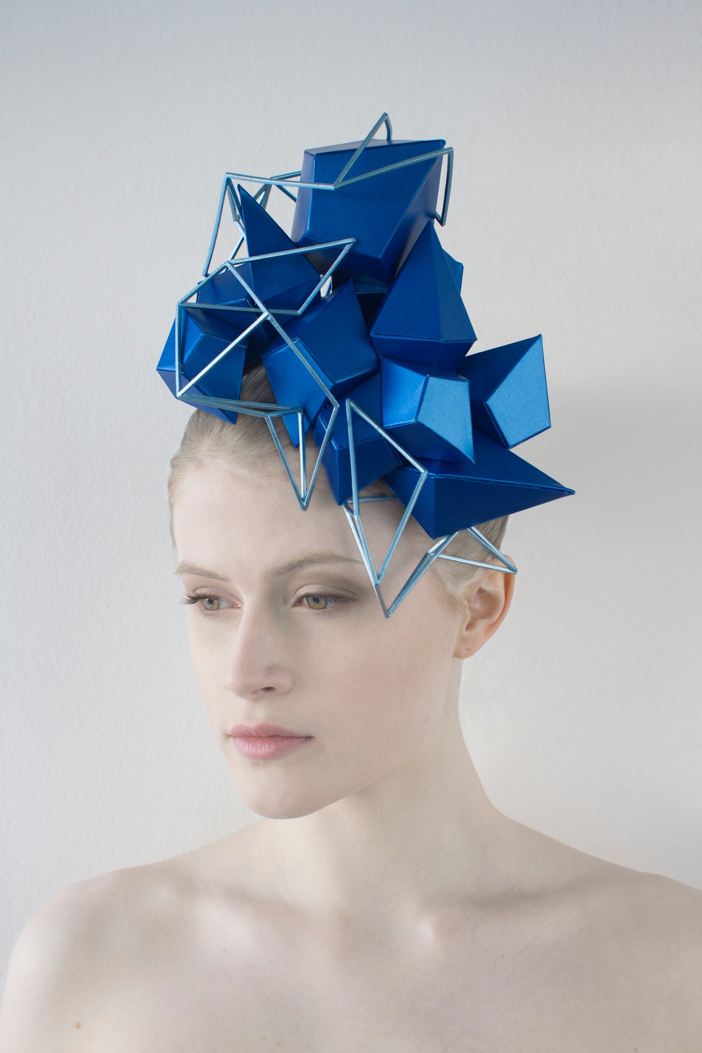 A woman sitting in front of a white background wears a shiny blue, geometric headpiece that is enhanced with a light-blue lattice. 