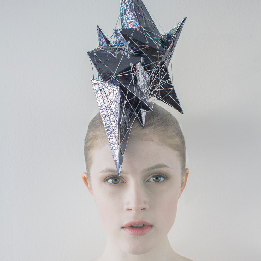 A woman in front of a white background wearing a headpiece made of shiny purple pyramids and silver thread. 
