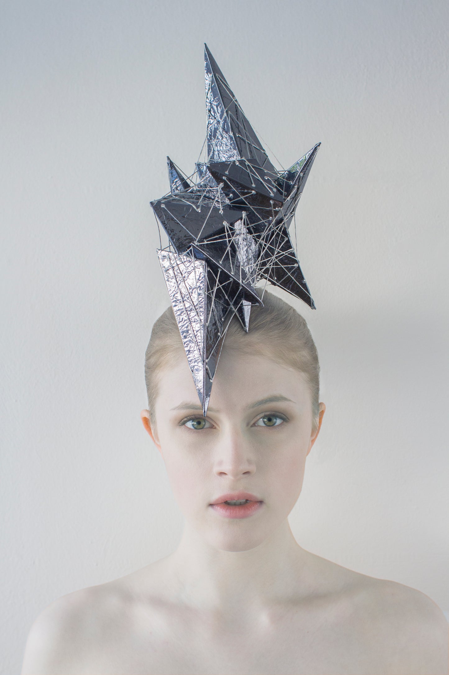 A woman in front of a white background wearing a headpiece made of shiny purple pyramids and silver thread. 