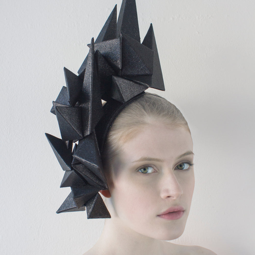 A headpiece made from black pyramids hugs the right side of a woman's head, as she sits in front of a white background. 