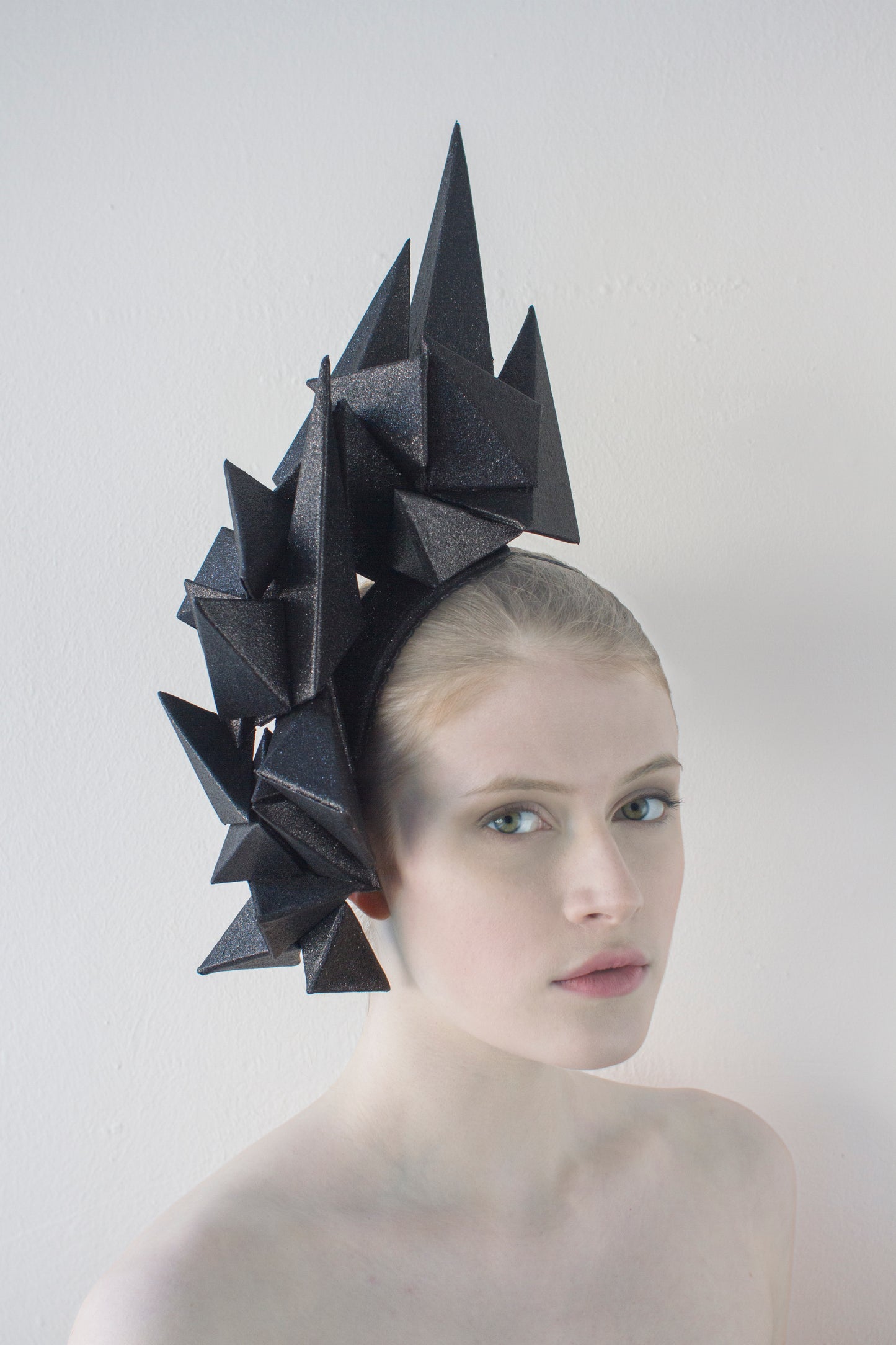 A headpiece made from black pyramids hugs the right side of a woman's head, as she sits in front of a white background. 