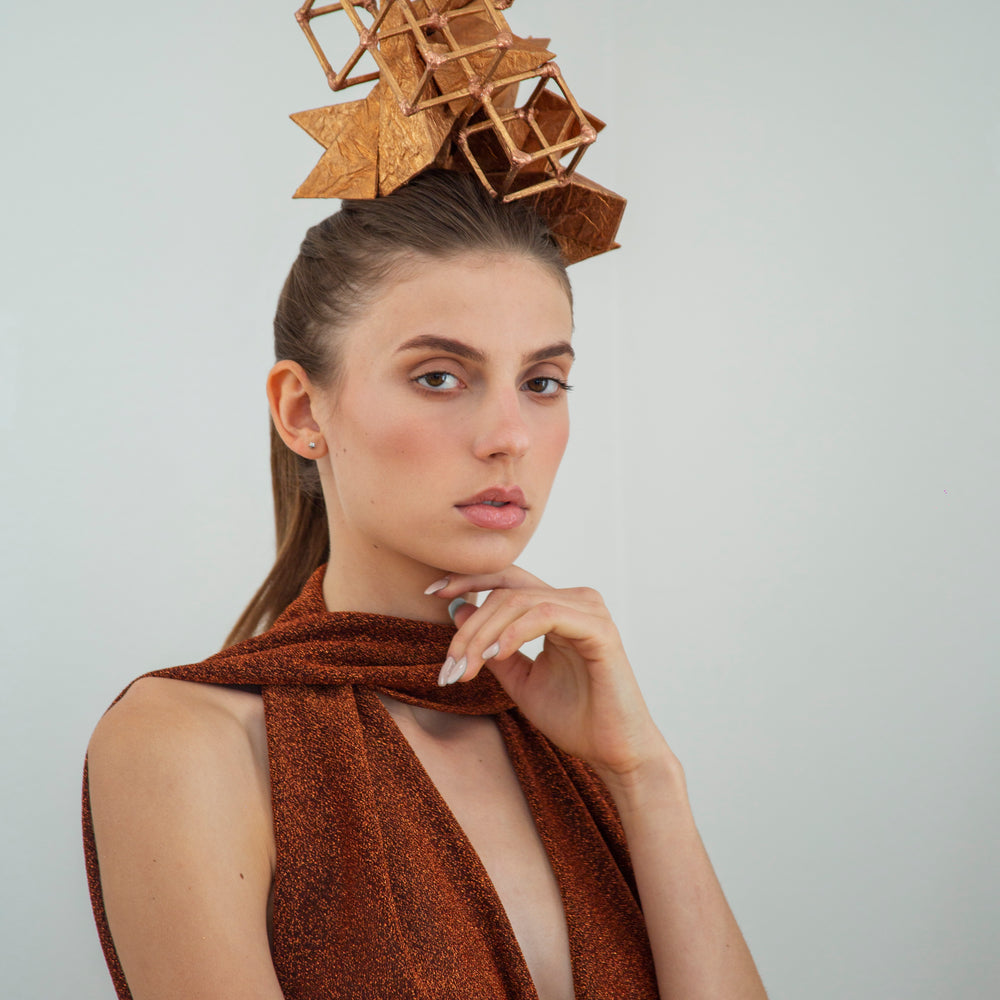 A woman is standing, touching her chin with her left hand and is wearing a copper-colored geometric headpiece made from pyramids and cubes. 