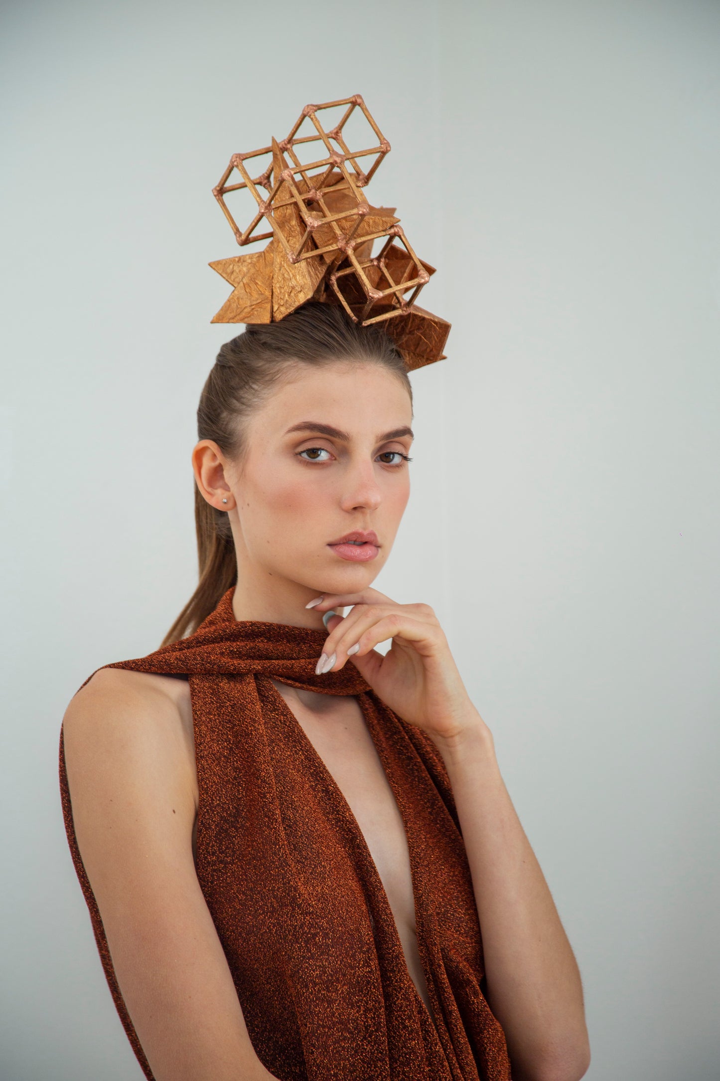 A woman is standing, touching her chin with her left hand and is wearing a copper-colored geometric headpiece made from pyramids and cubes. 