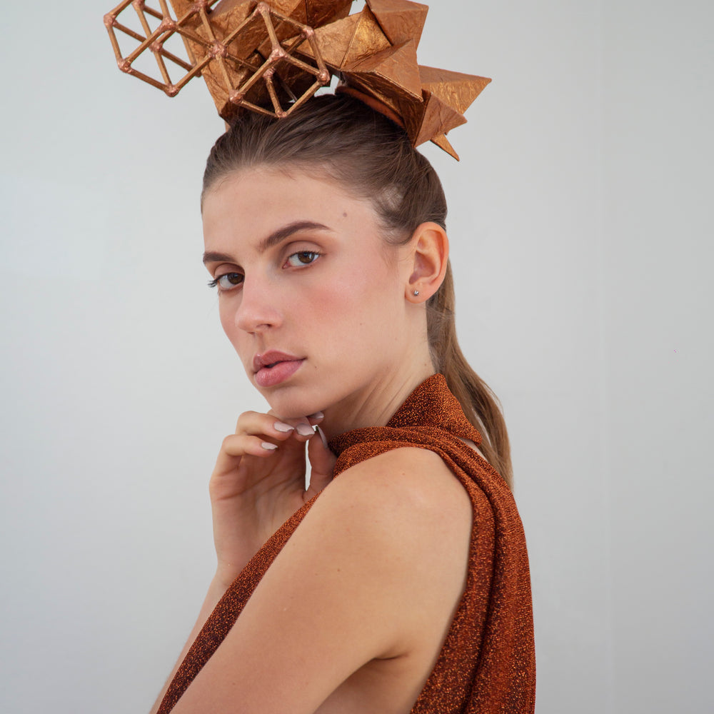 
                  
                    A woman in a copper-colored shirt looks to her left, touching her chin with her right hand and wears a copper-colored geometric headpiece made from pyramids and cubes. 
                  
                